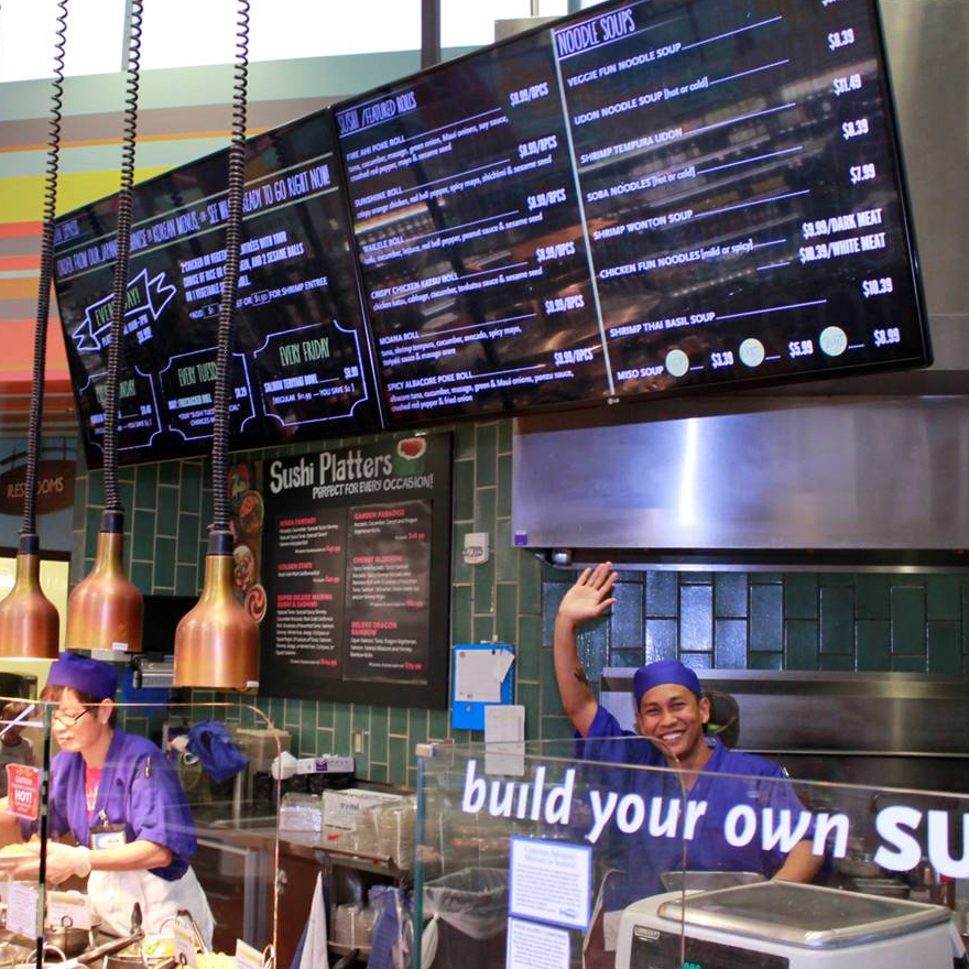 A smiling kitchen worker stands below two large video menus and waves from behind a glass barrier that says "Build your own sushi."