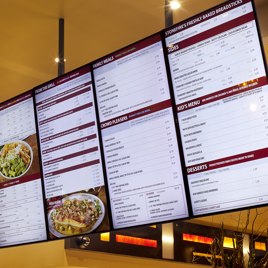 Six vertical digital menu boards are hung from a restaurant ceiling.