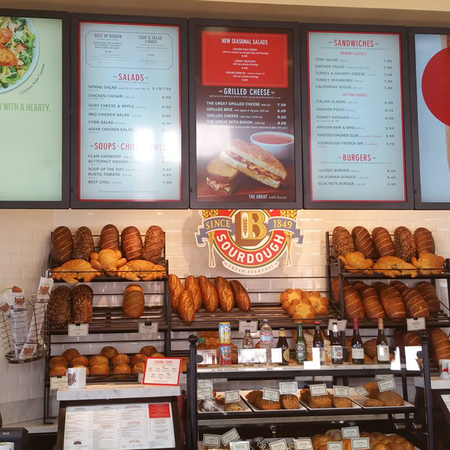A tall rack of baked bread loaves underneath five vertical video menu screens showing soups and sandwiches.