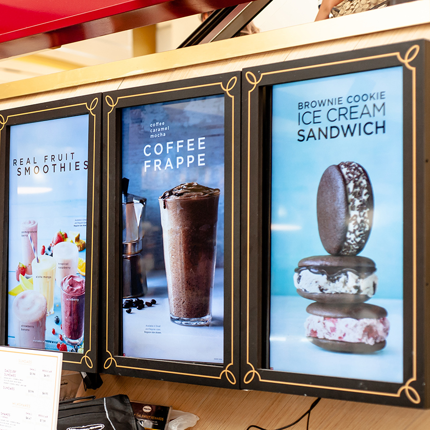 Three vertical digital signs showcase blended coffee and smoothies behind a counter holding ingredients and tools.