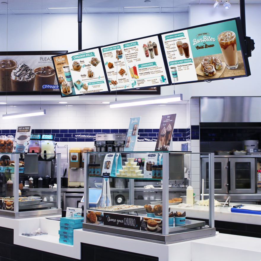 A brightly lit Cinnabon storefront showing white countertops, two glass display cases, and three digital menus hung overhead.
