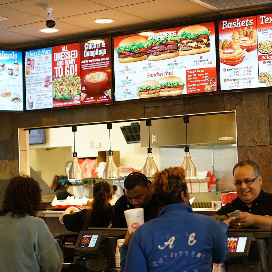 Customers and employees are gathered around a QSR counter as digital menus display food items overhead.