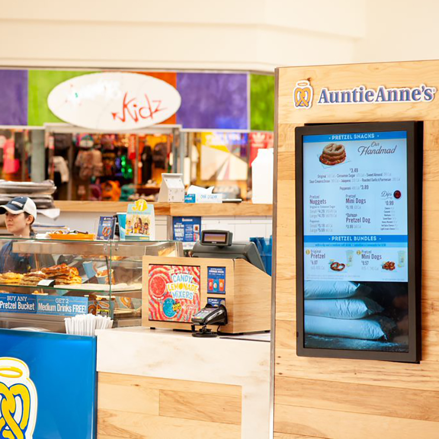 Two people are ordering at an Auntie Anne's counter as a vertical digital menu is in the foreground.