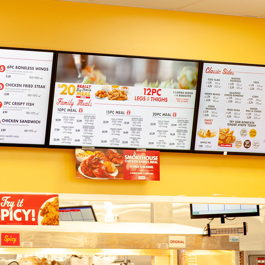 A row of horizontal digital menu boards showcase fried chicken and sides above a fast food heating shelf.