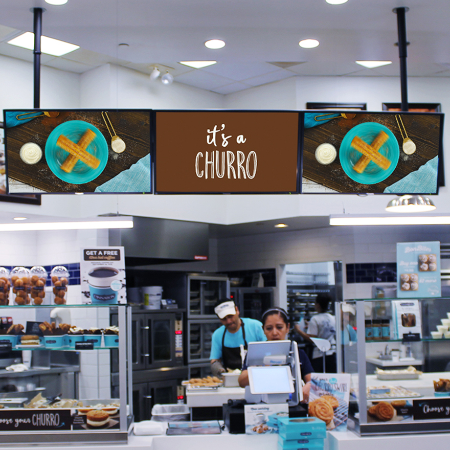 Three video menu boards advertise churros above a Cinnabon front counter.