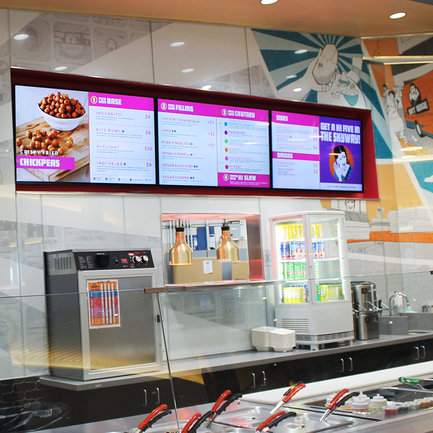 A stainless steel quick-service restaurant counter lined with bins of ingredients with three horizontal digital menus in the background.