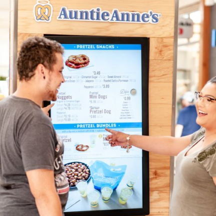 Two people are smiling and pointing toward menu items on a digital menu board for Auntie Anne's pretzels. 							