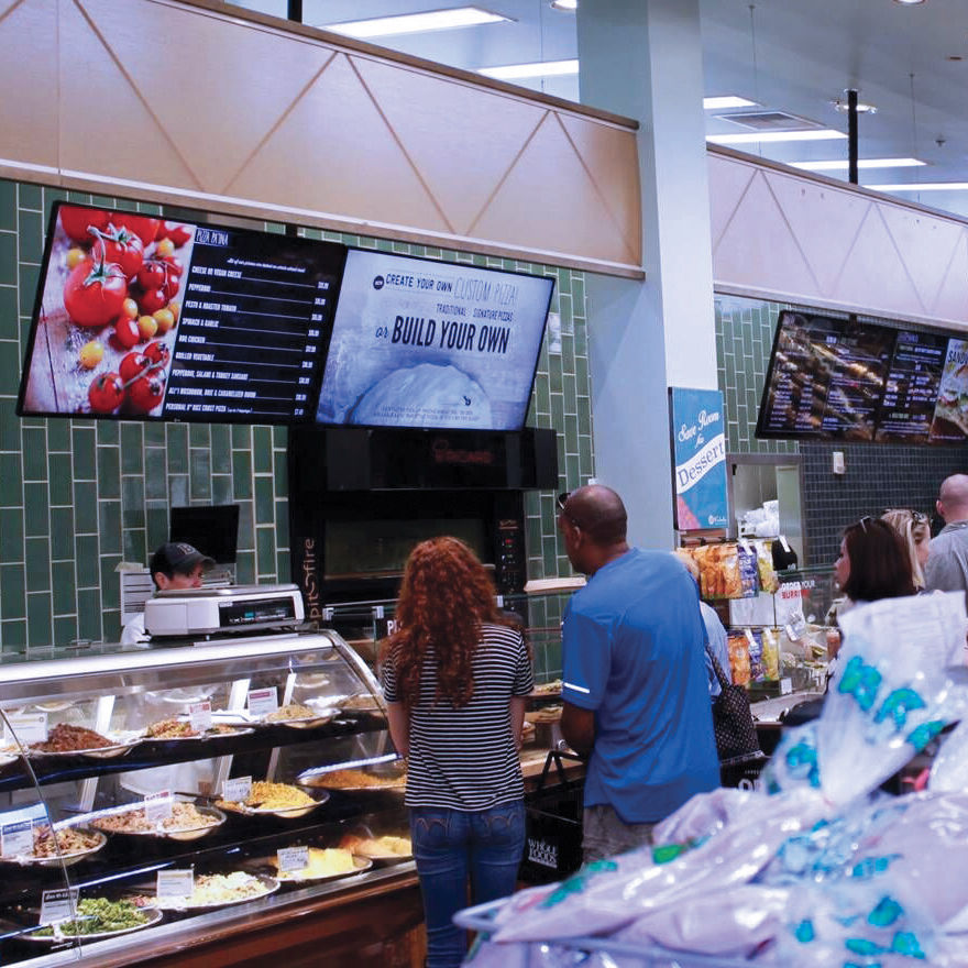 Six shoppers are browsing a deli counter while four horizontal menu boards hang overhead.
